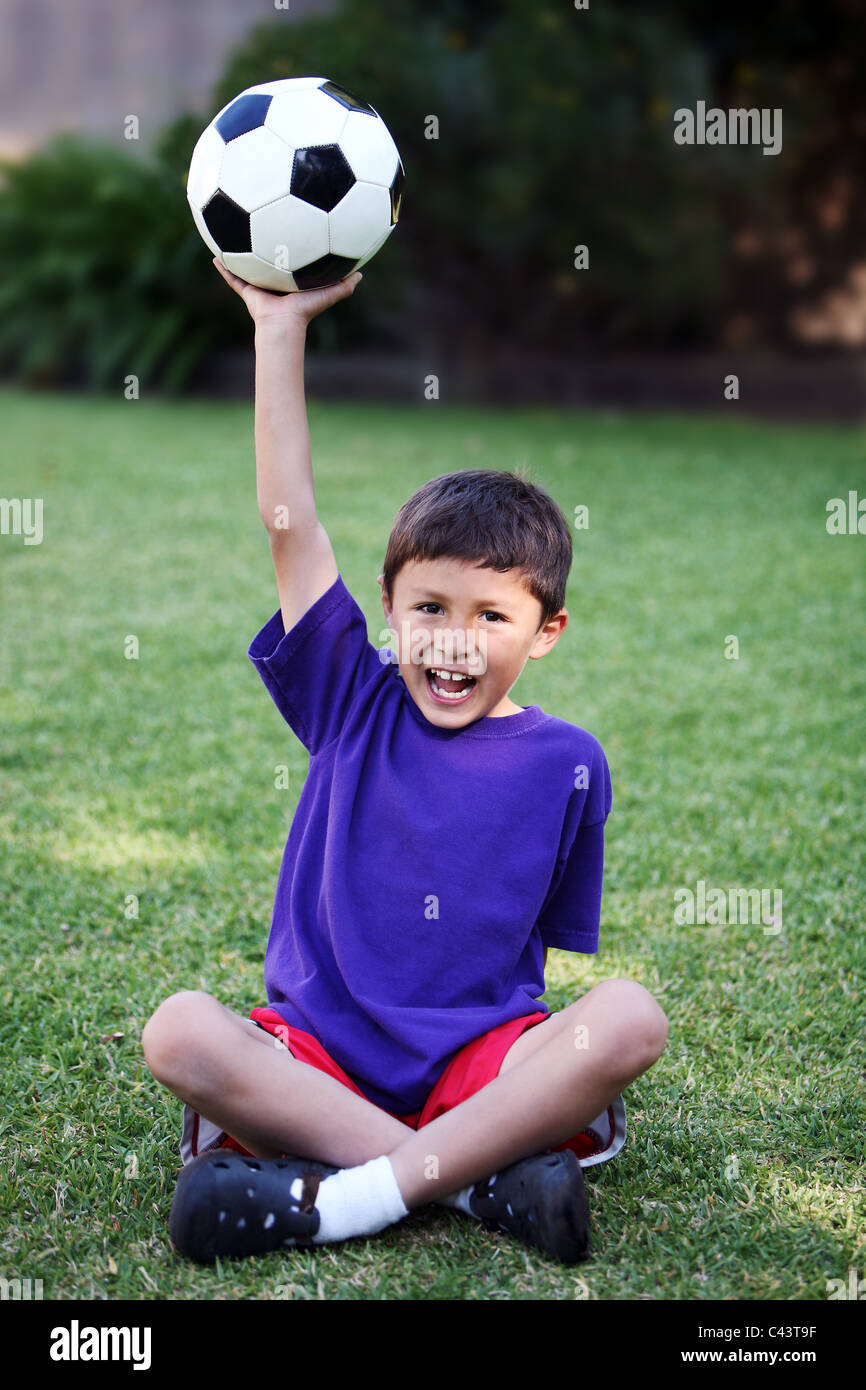 Young boy holds up soccer ball while sitting on grass Stock Photo