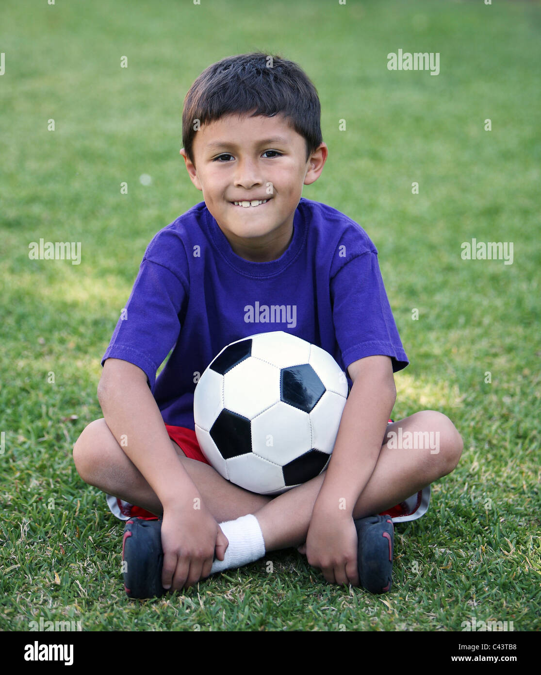 Authentic young Latino boy sitting cross-legged with soccer ball on field of grass Stock Photo