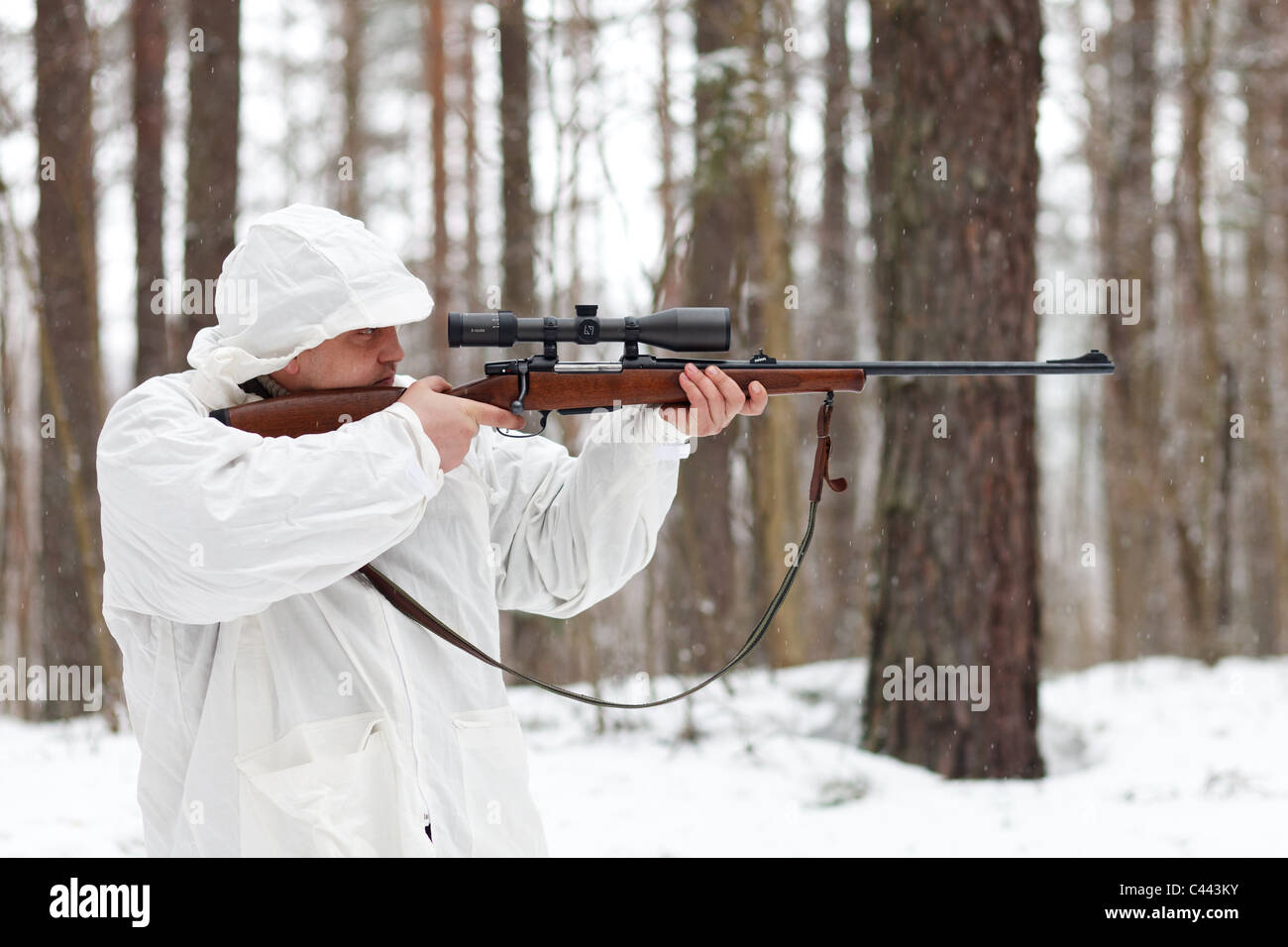 Sniper in white camouflage aiming with rifle at winter forest. Stock Photo