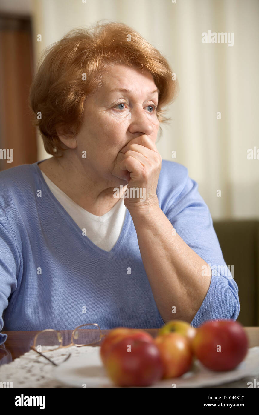 A worried senior woman sitting at a table with her hand covering her mouth Stock Photo