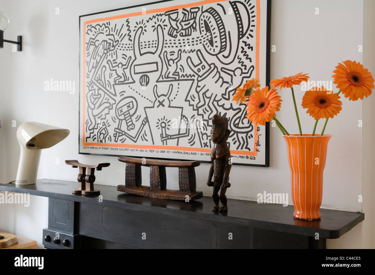 Smaller Tsonga and larger Zulu headrest on mantelpiece with Keith Haring artwork and orange vase with gerbera Stock Photo