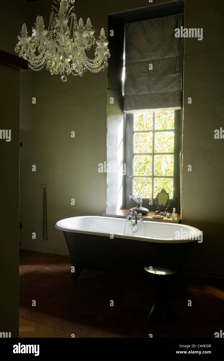Freestanding roll-top bath in bathroom with chandelier and window with blind Stock Photo
