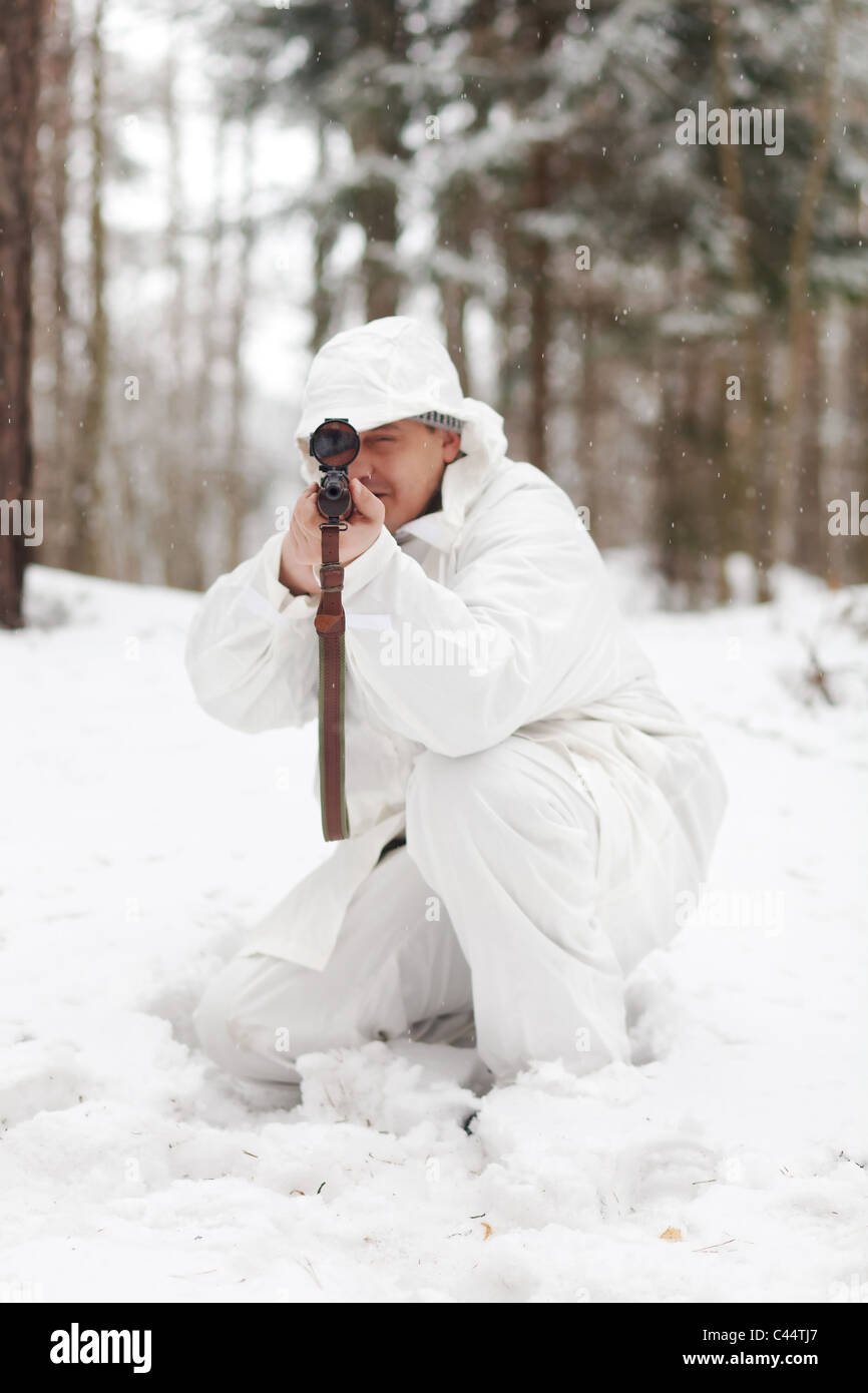 Soldier in white camouflage aiming with sniper rifle at winter forest. Stock Photo