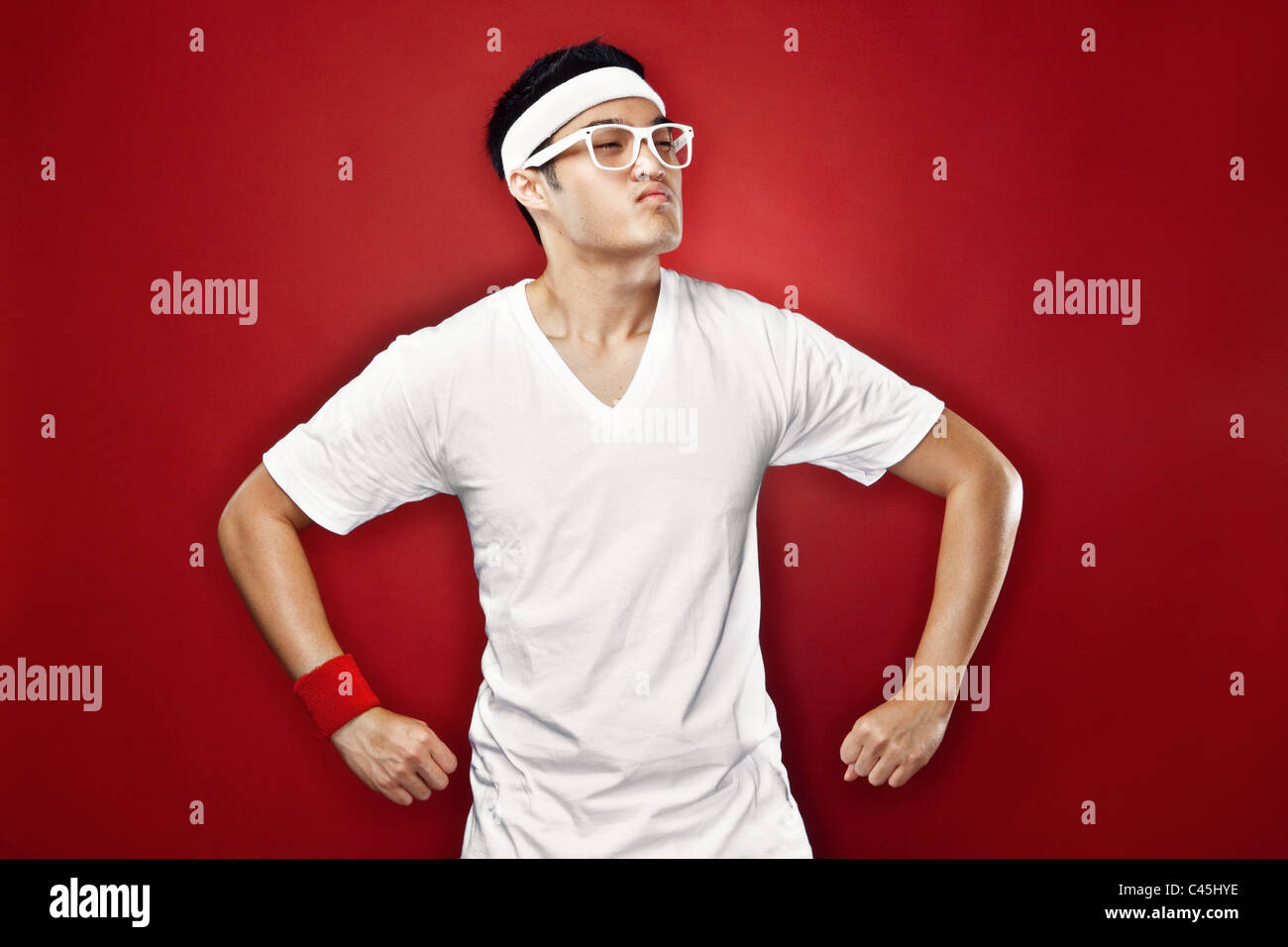 Studio portrait of Asian male teenager doing a superhero pose in white athletic gear & nerdy glasses against a red backdrop. Stock Photo