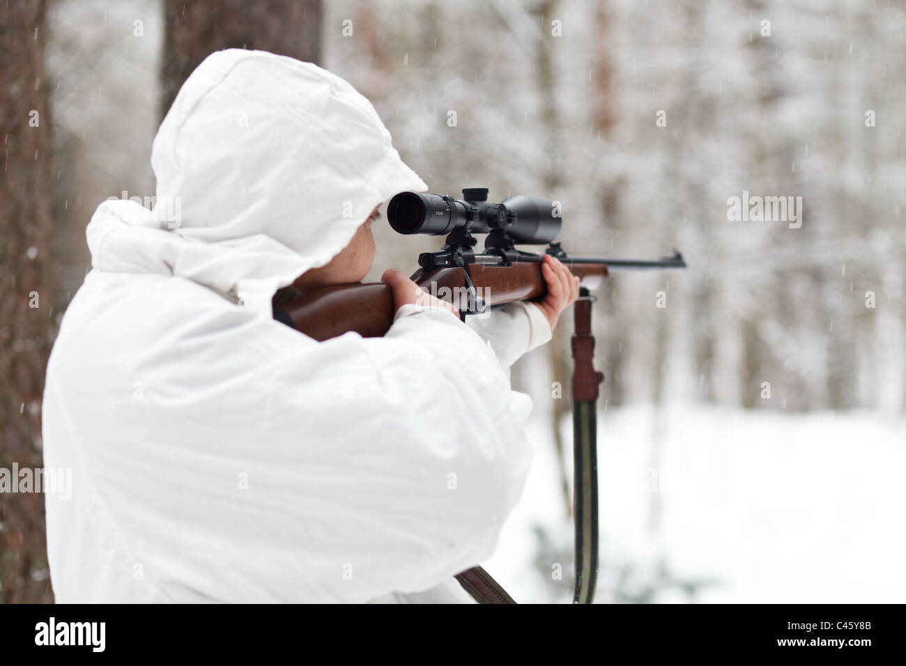 Soldier in white camouflage aiming with sniper rifle at winter forest. Stock Photo