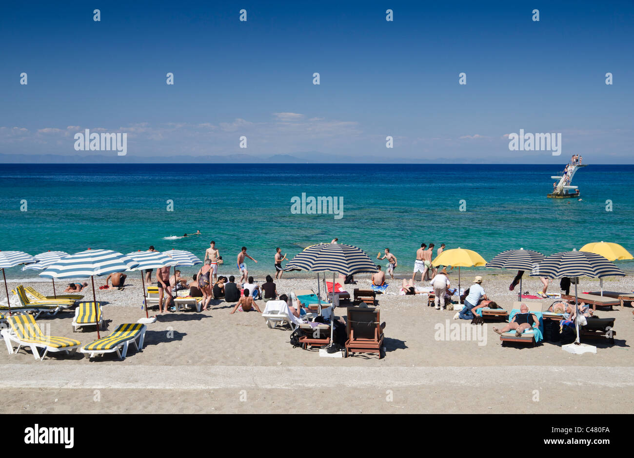 People on a sandy beach in Rhodes Town, with a diving board in the sea