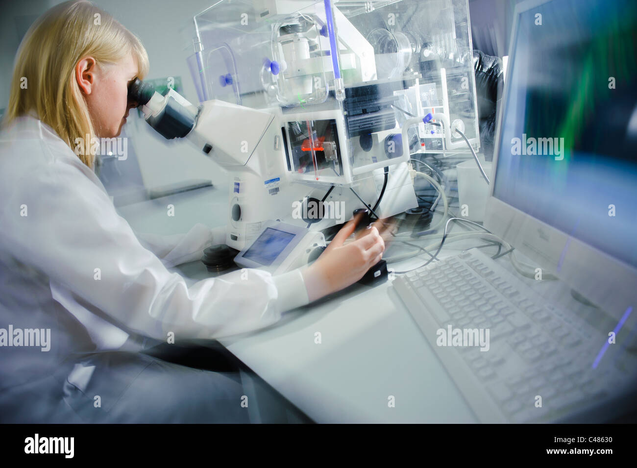 Young blond female scientist white lab coat powerful microscope in well lit science laboratory cell images on computer screen Stock Photo