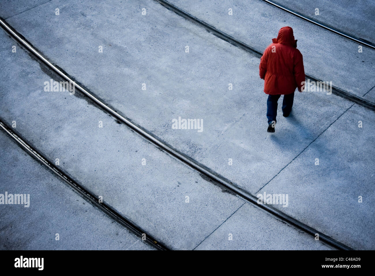 A man wearing a red coat with a hood walks forlornly over tram lines in Manchester. Stock Photo