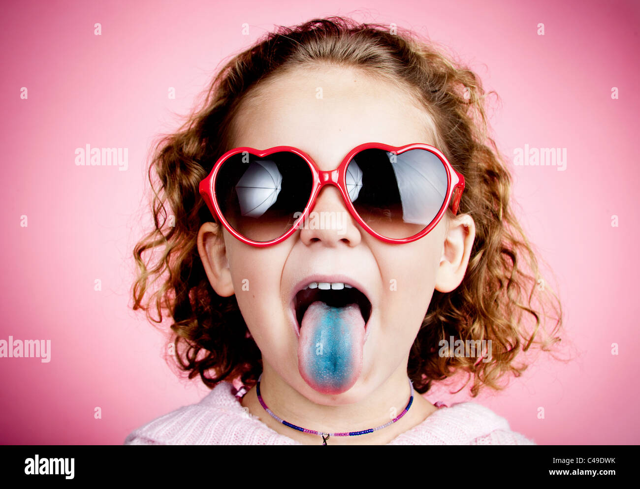 A young girl with curly hair on a pink studio wall wearing heart-shaped sunglasses and sticking out a stained blue tongue Stock Photo