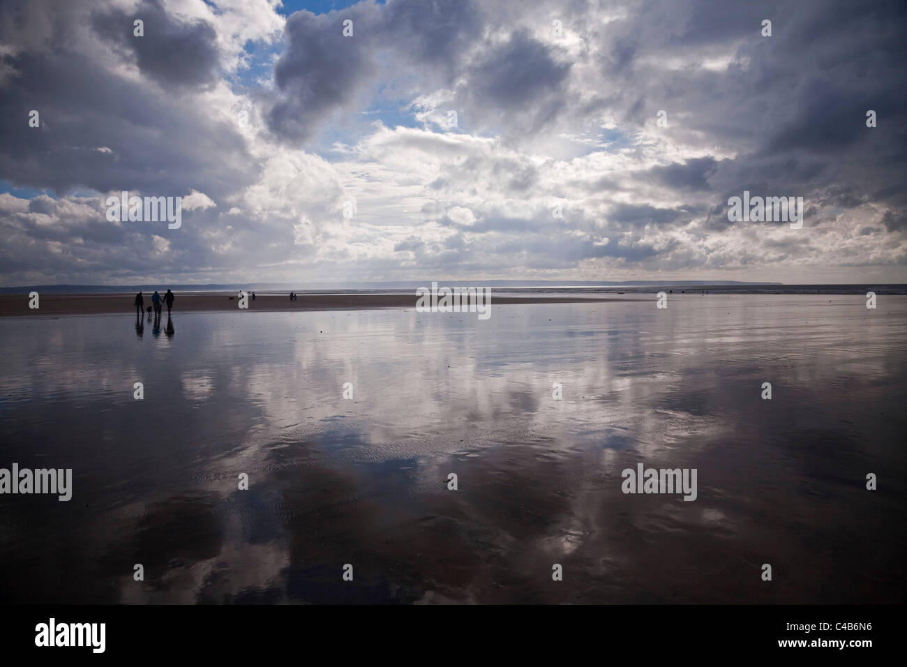 UK, Devon. A group of friends walk across the beach at Saunton Sands, with the gathering clouds reflected at low tide. Stock Photo