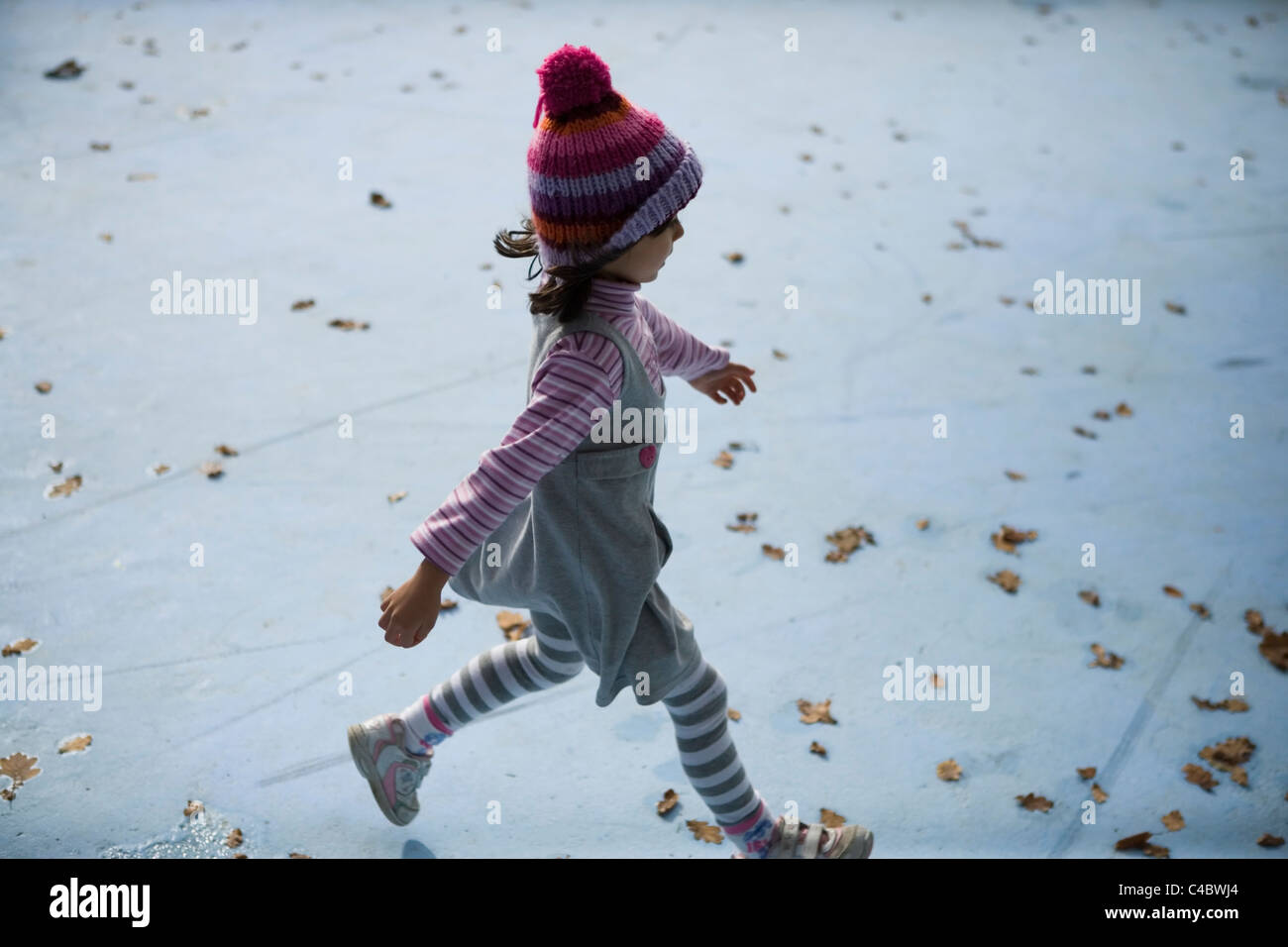 Girl aged four with knitted woollen bobble hat running through empty blue paddling pool filled with autumnal leaves Stock Photo