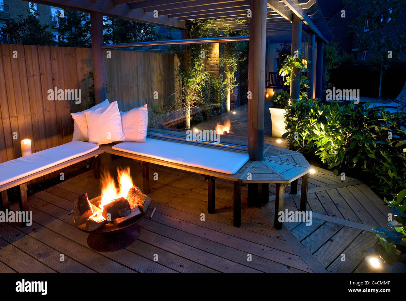 contemporary modern English town garden with wooden bench decking and pergola, glass wind break and fire basket at night Stock Photo