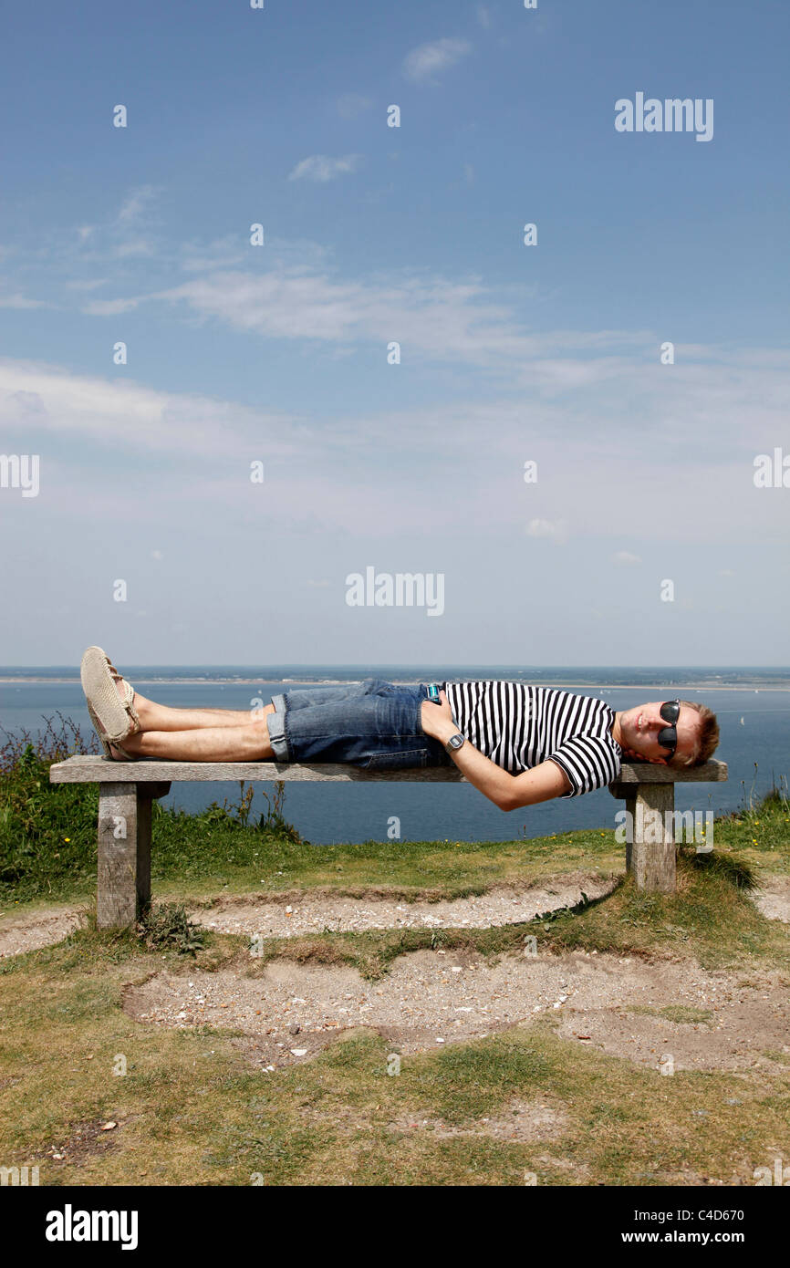 Man wearing striped T shirt on holiday lying on a cliff top bench relaxing and having a rest - Model Released Stock Photo