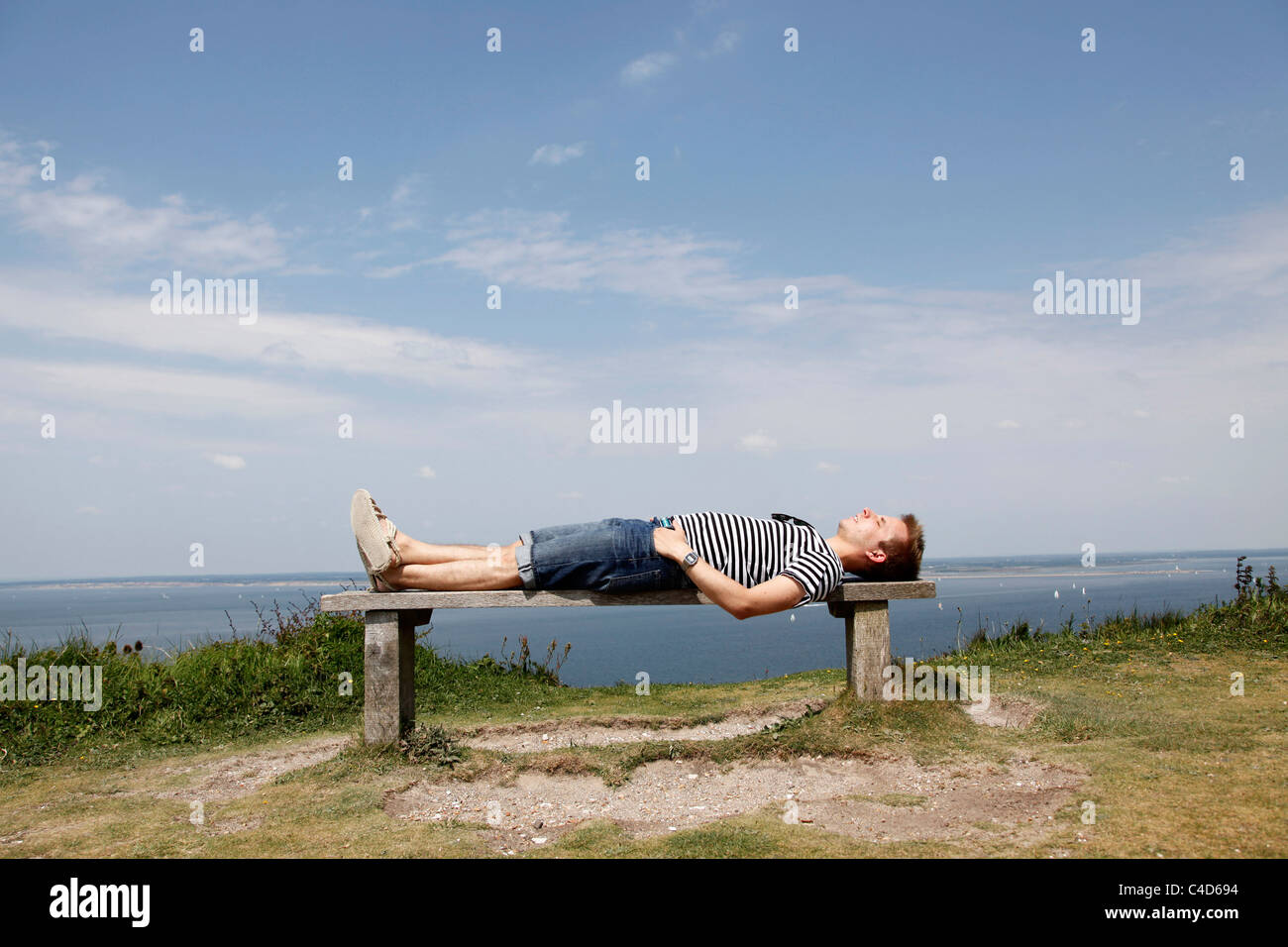 Man wearing striped T shirt on holiday lying on a cliff top bench relaxing and having a rest - Model Released Stock Photo