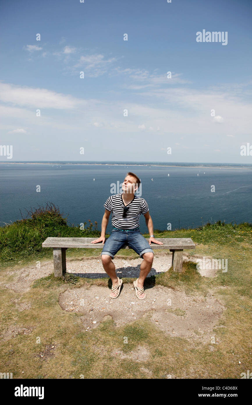 Man wearing striped T shirt on holiday sitting on a cliff top bench relaxing and having a rest enjoying the sun - Model Released Stock Photo