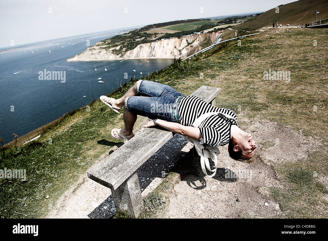 Man wearing striped T shirt on holiday leaning back on a cliff top bench having fun and stretching - Model Released Stock Photo