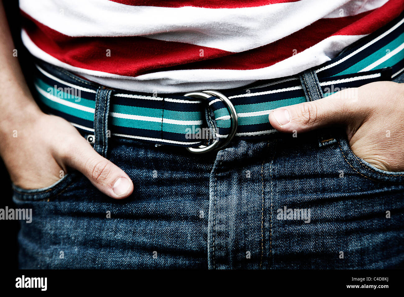 Man with his hands in his pockets wearing blue denim jeans trousers with a green, blue and white fashion belt Stock Photo