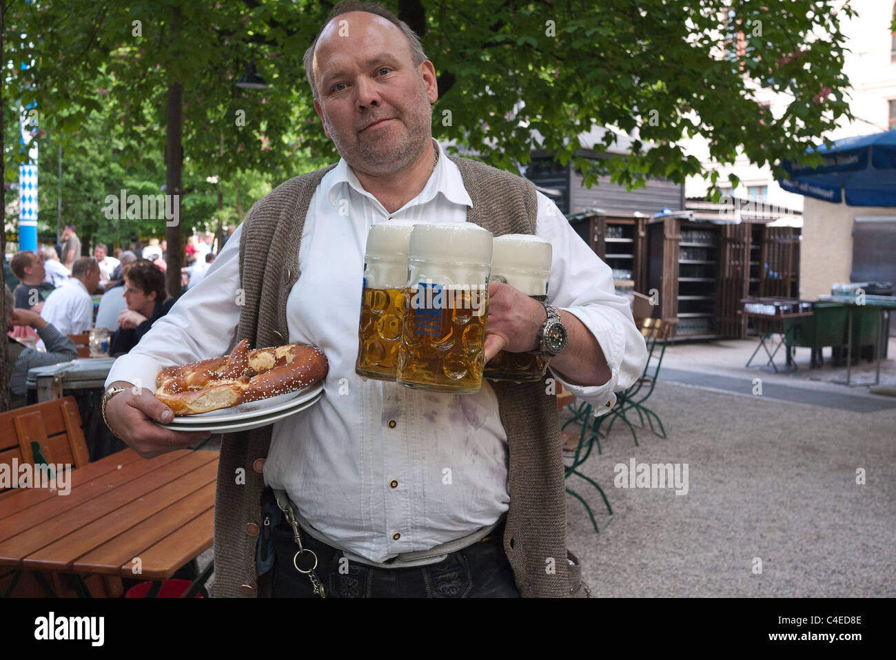 A waiter carries three very large beer steins & one of the traditional over-sized pretzels at the Augustiner-Keller beer garden. Stock Photo