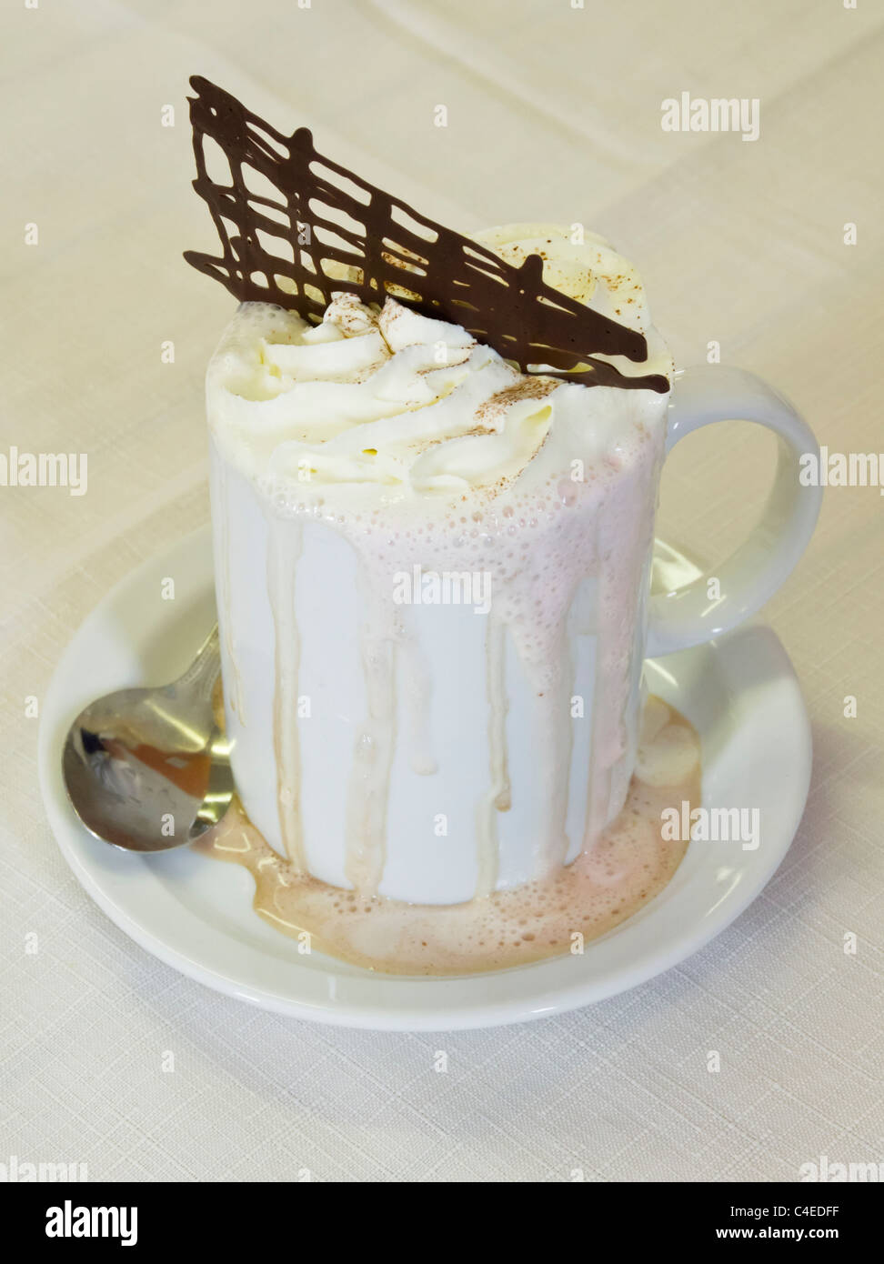 Mug of frothy hot chocolate drink topped with whipped cream running down side into the saucer on a white tablecloth Stock Photo