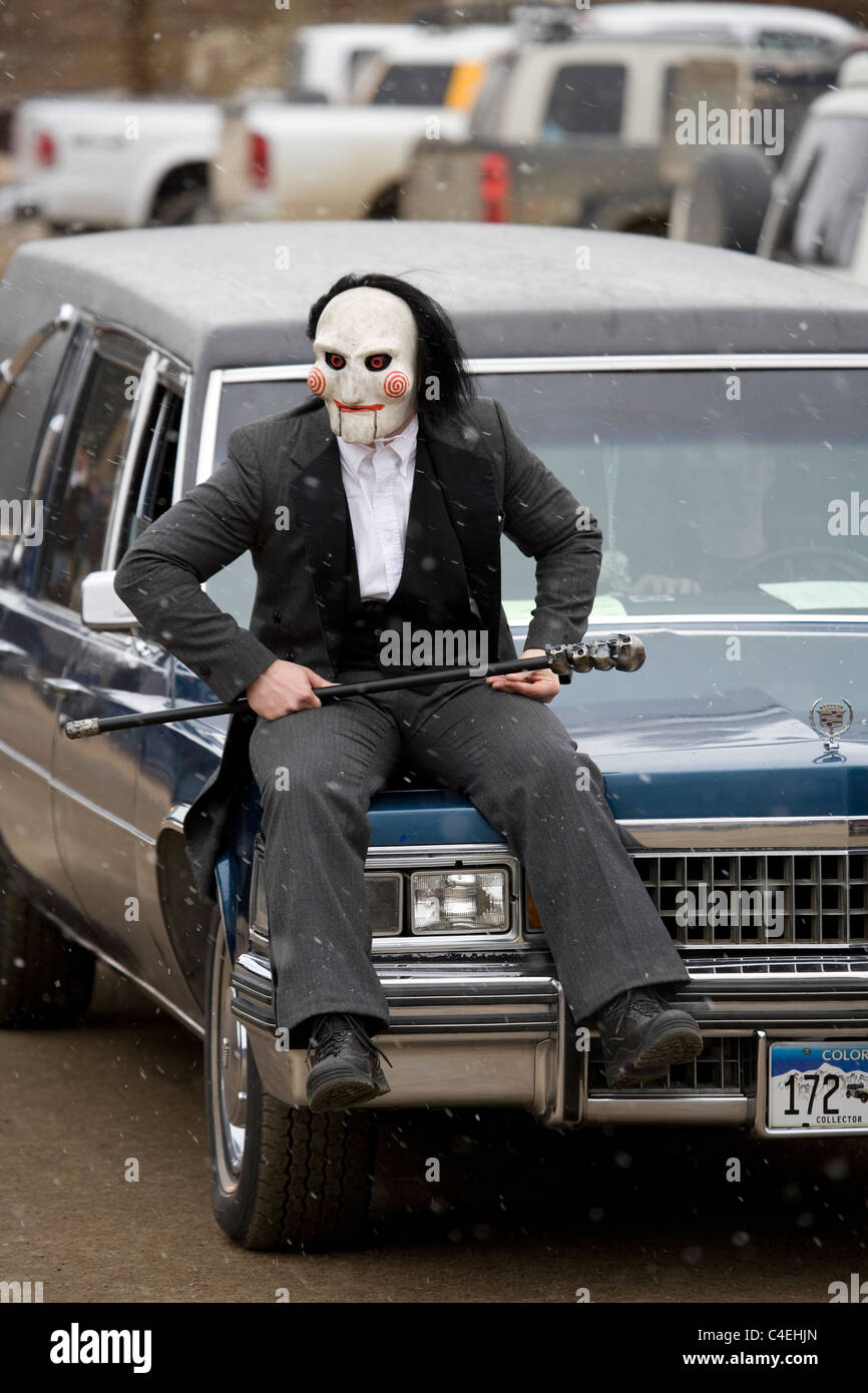 A person dressed in a ghoul costume rides on the hood of a hearse