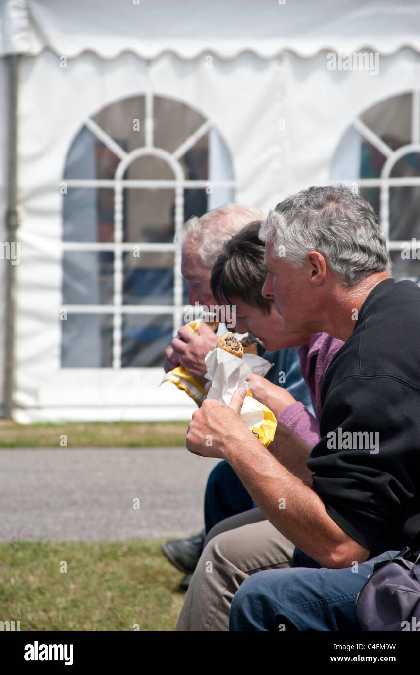 Three people sitting side by side on a bench, eating Cornish pasties from paper bags. Behind them is a white tented pavillion Stock Photo