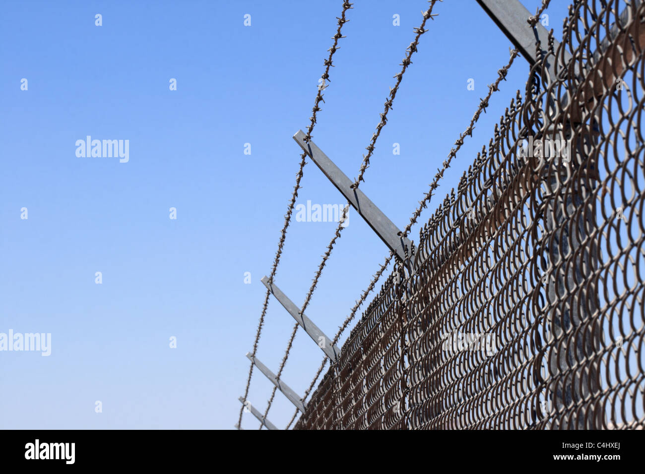 horizontal image of a chain link security fence topped with three strands of barbed wire and blue sky background Stock Photo