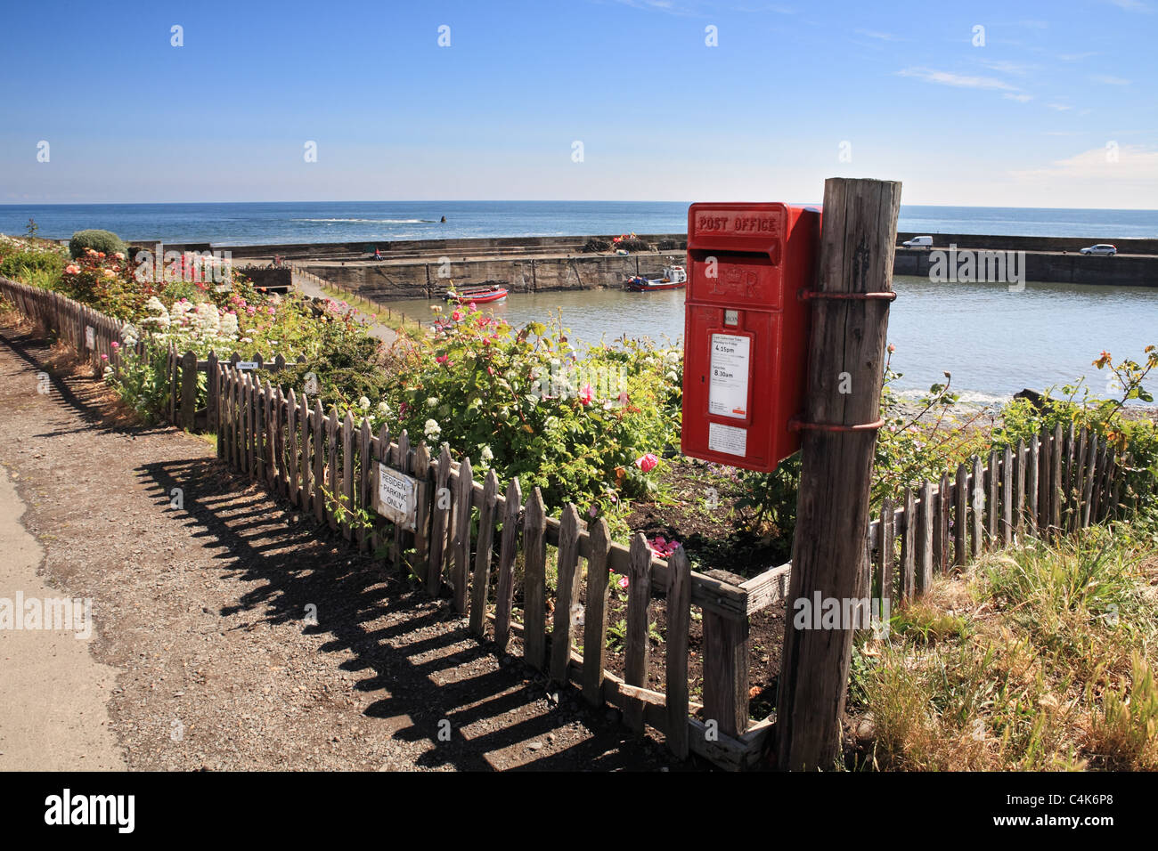A red post box with Craster Harbour in the background. Northumberland, North EastEngland, UK Stock Photo