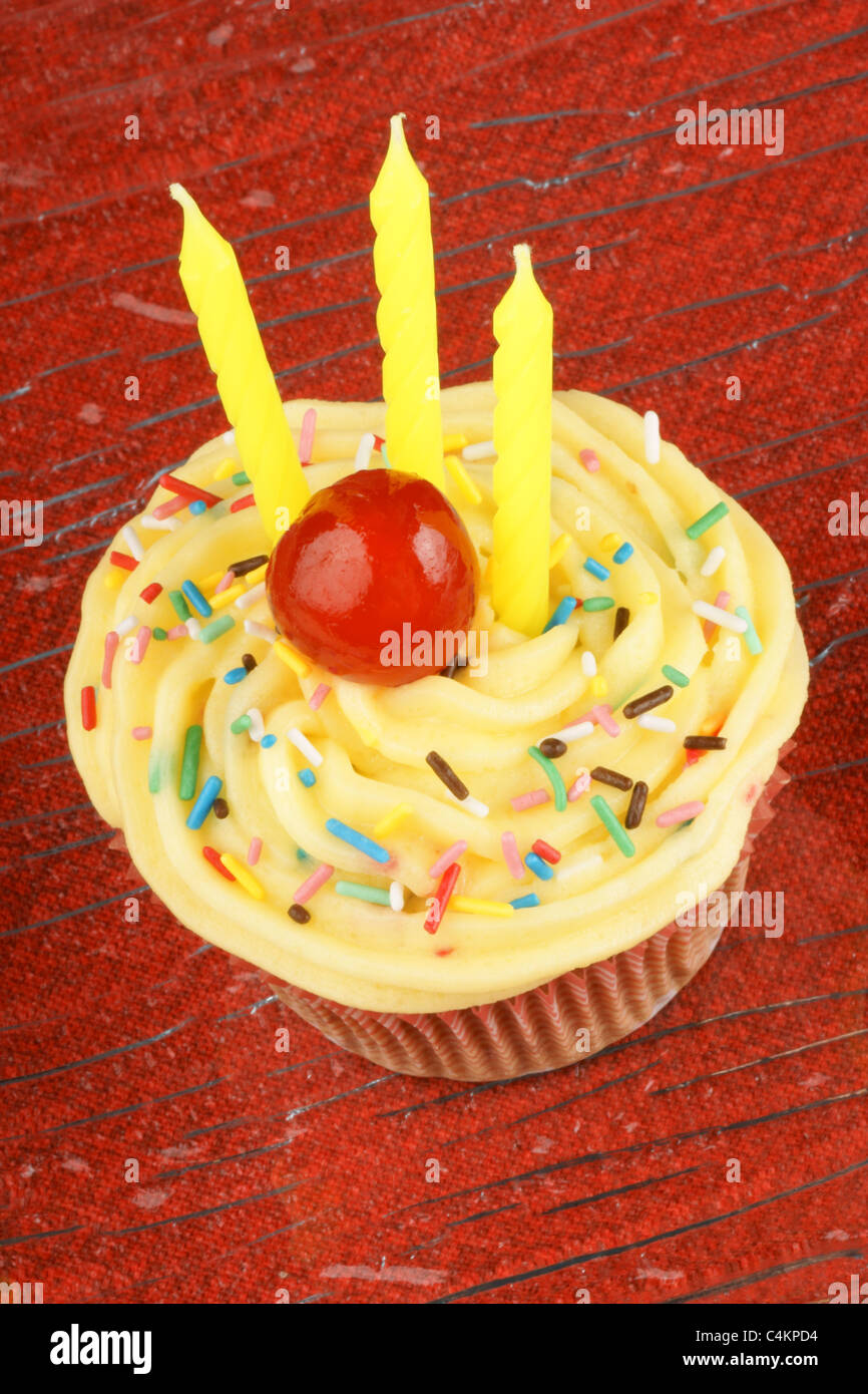 Birthday cupcake with lemon butter-cream, sprinkles, candied cherry and three candles. Over a red background. Selective focus. Stock Photo