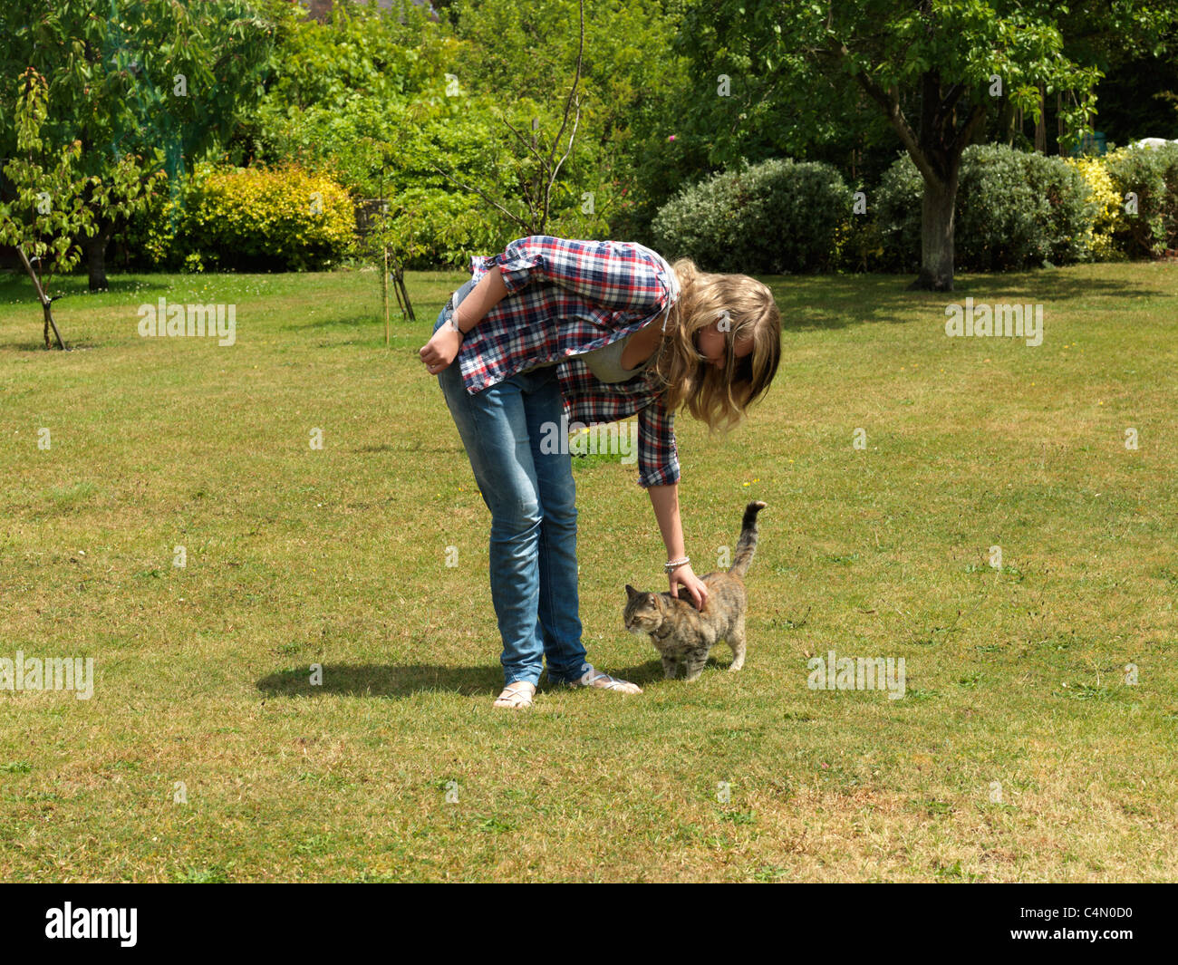 Young Woman Out In The Garden Wearing Shirt, Jeans And Sandals Stroking A Tabby Cat Stock Photo