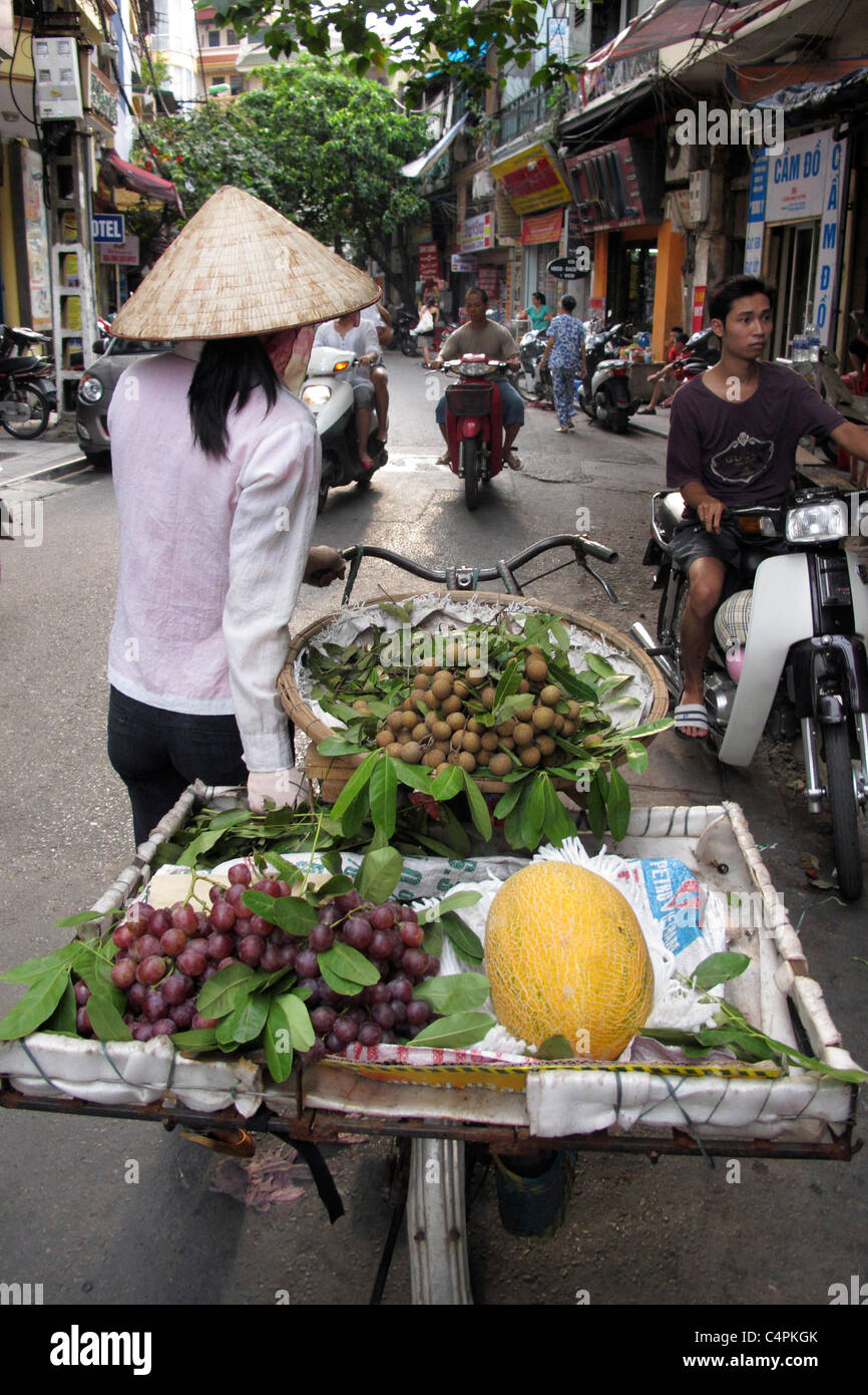 A Vietnamese woman with a typical conical hat pushes a motor bike carrying fruits (melon and grapes) on Hanoi streets Stock Photo