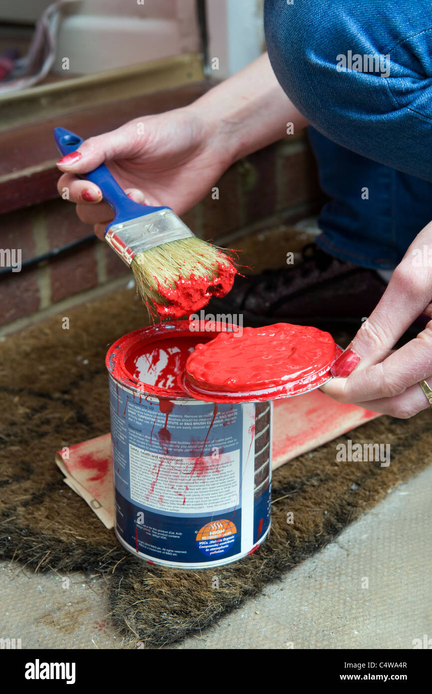 Close up of young Caucasian womans hand holding a dripping paint brush loaded with red paint over a paint pot Stock Photo