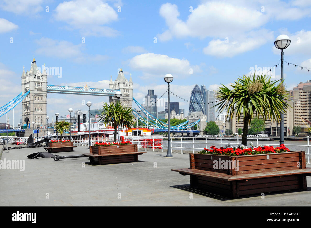 Butlers Wharf Thames Path promenade beside River Thames views of Tower Bridge & City of London skyline row cordyline trees red flowers in planters UK Stock Photo