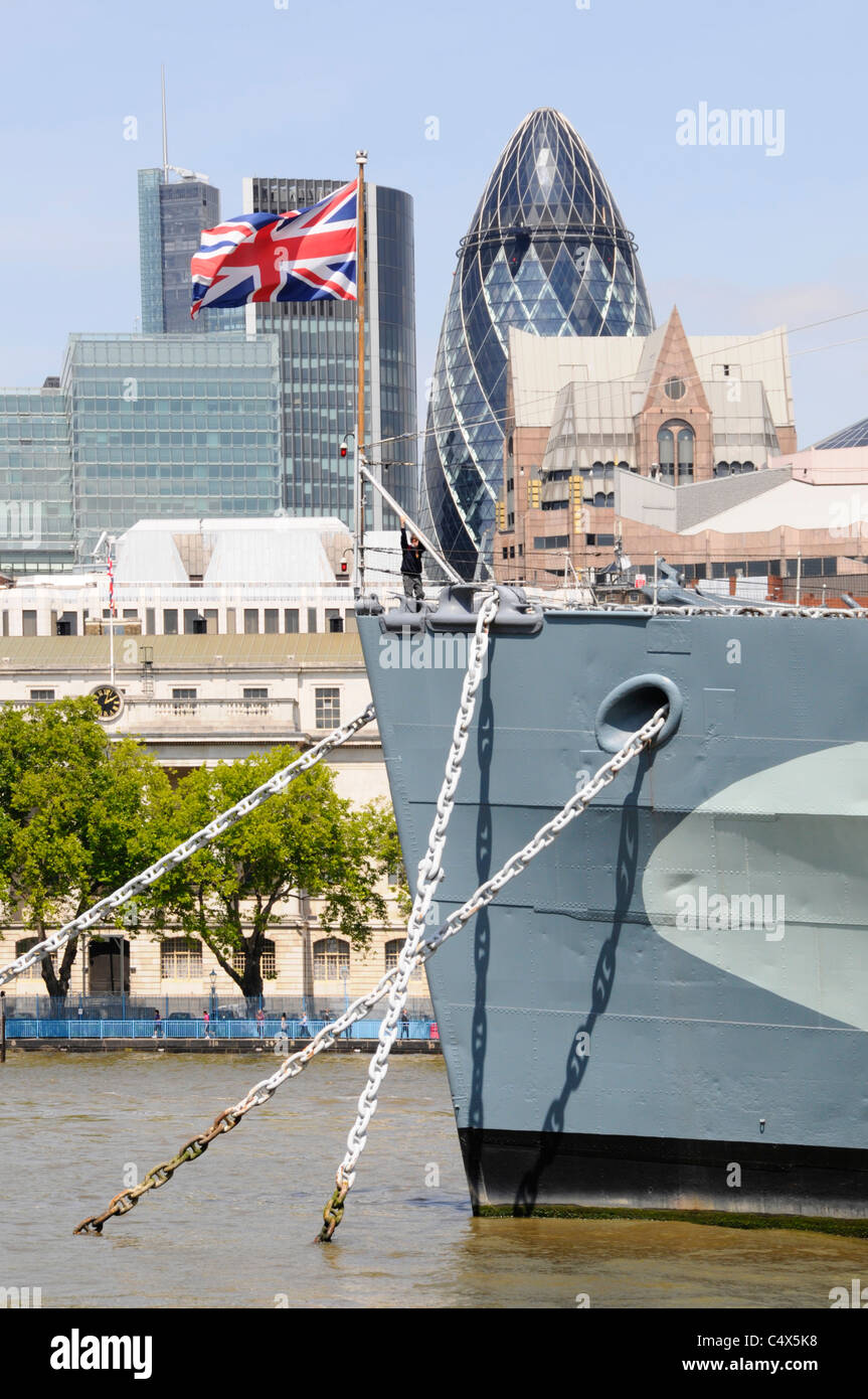 Union Jack flying on bow of historical HMS Belfast museum ship with City of London skyline of skyscraper landmark business office blocks England UK Stock Photo