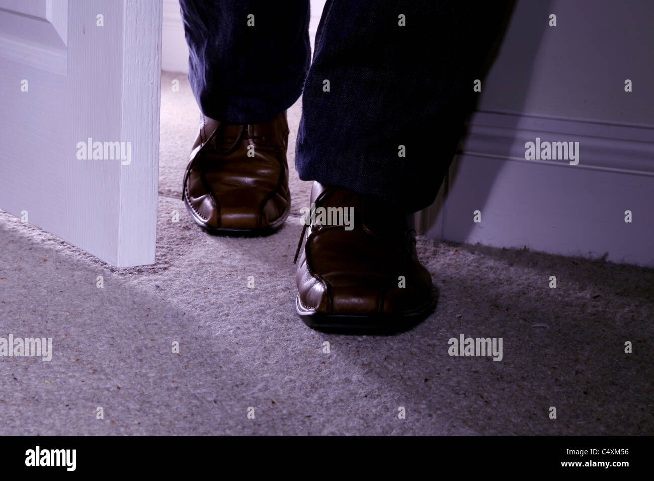 Man's feet only wearing brown shoes entering a dark carpeted room Stock Photo