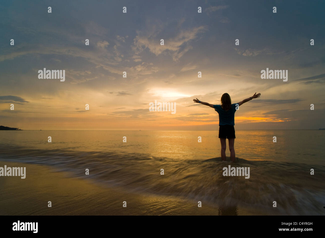A young woman at the beach, standing in the sea at sunset with her hands in the air, celebrating freedom or worshipping God Stock Photo