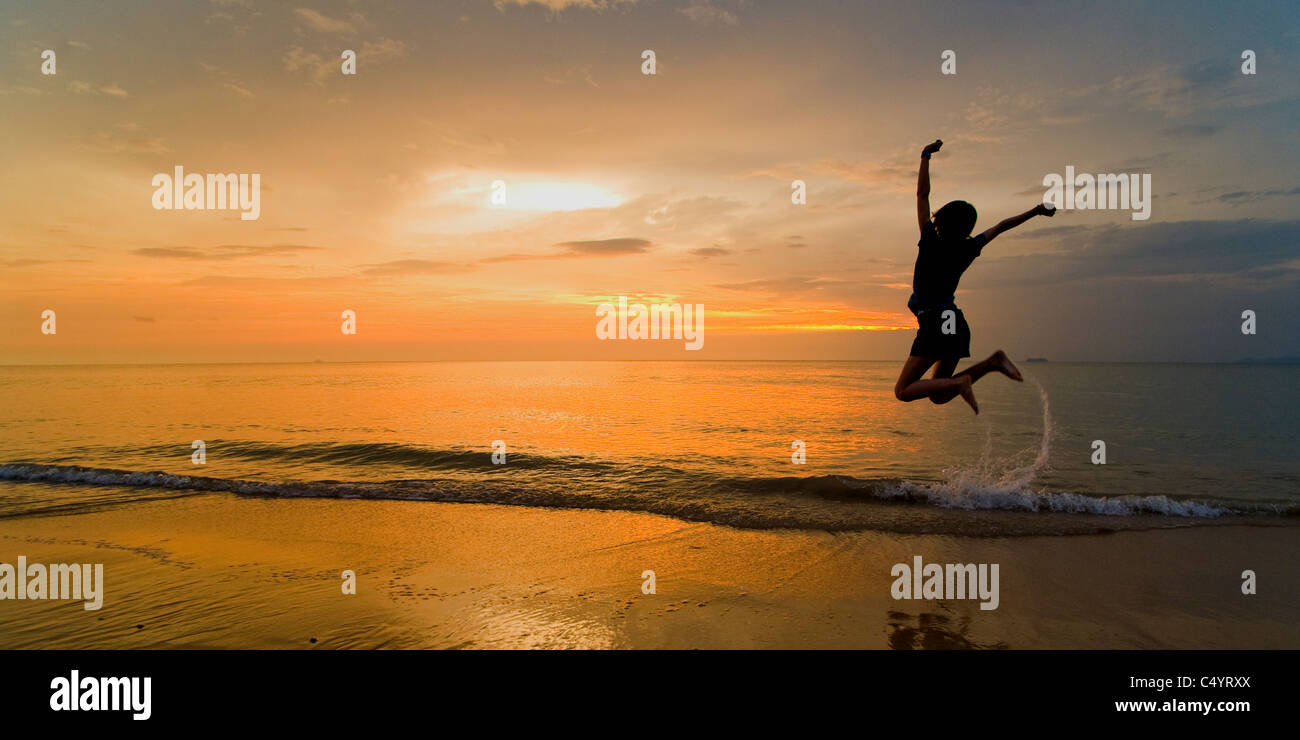 A panoramic photo of a young woman jumping for joy and celebrating on Phra Ae beach, Koh Lanta, South Thailand at sunset. Stock Photo