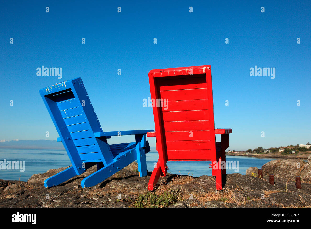 Red and Blue chairs sitting on rocks looking out to the pacific ocean on a calm sunny summer morning in Victoria BC Canada Stock Photo
