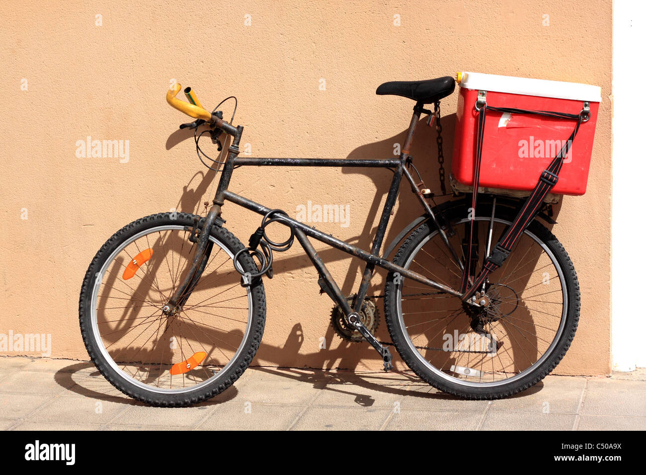 A rusty old black bike leaning against an ochre coloured wall.  A cool box is strapped to a rack at the back of the bicycle. Stock Photo