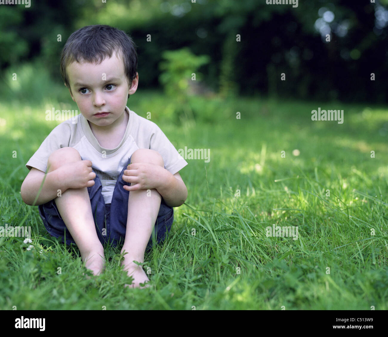 Little boy sitting in grass, looking away in thought Stock Photo