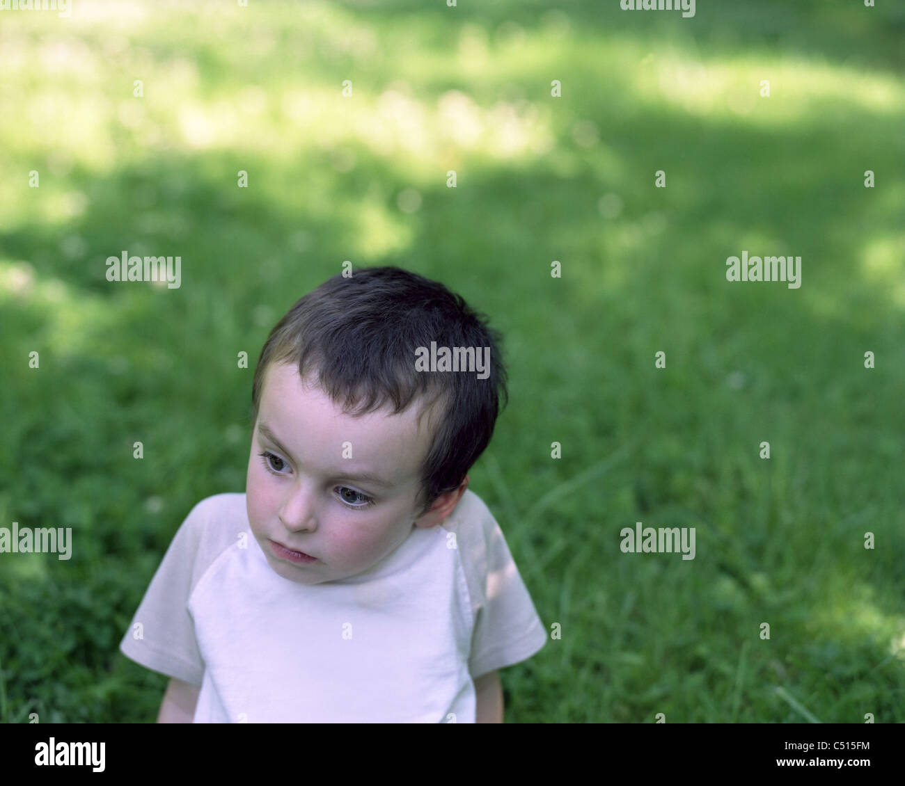 Little boy sitting in grass, looking away in thought Stock Photo