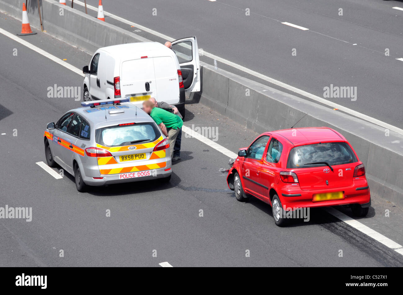 Aerial view UK motorway vehicles stationary in lane 4 accident involving red car below bridge witnesses talk to police beside concrete crash barrier Stock Photo