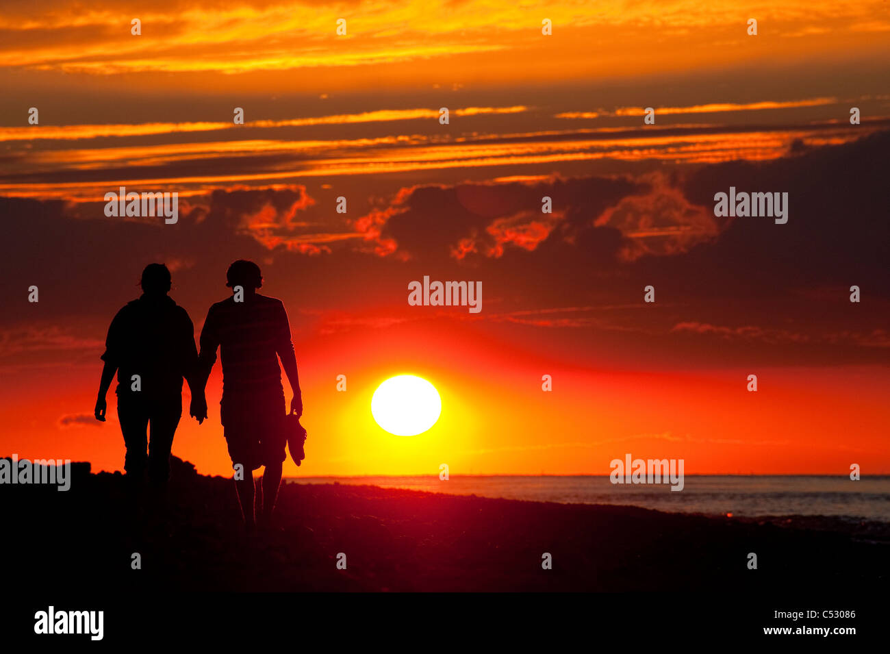 Couple walking hand in hand along a beach at sunset, on the Norfolk ...