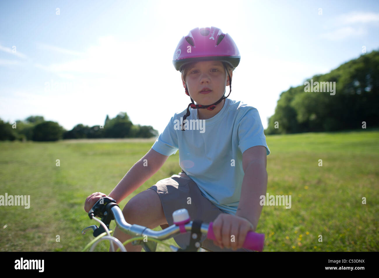 A young girl on a bicycle wearing a pink helmet photographed into the sun to induce lens flare for effect. Looking into camera. Stock Photo