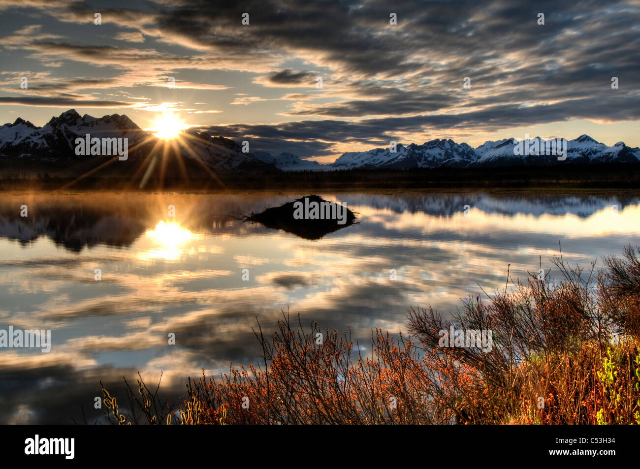 Sun rises over the Chugach Mountains with a pond and beaver lodge in the foreground, Chugach National Forest, Alaska Stock Photo