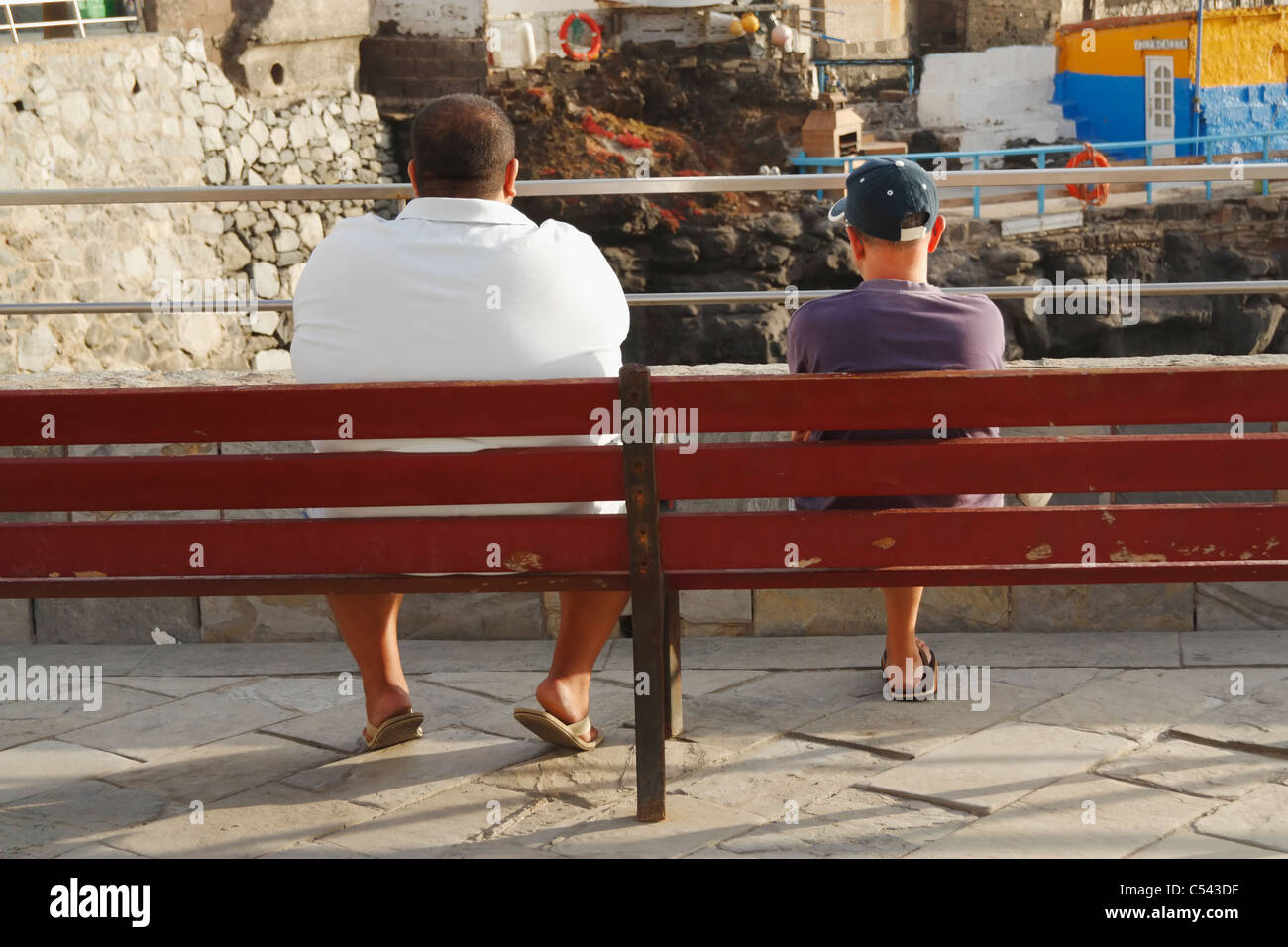 Large Man And Small Man Sitting On Bench In Spain Stock Photo