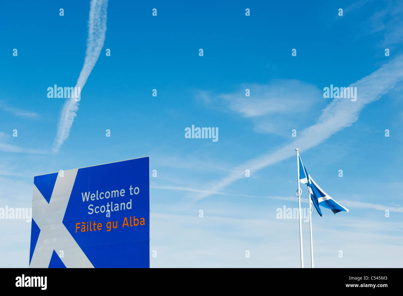 Scotland border sign at the Northumberland border in front of a white cross plane trail in the blue sky and scottish flags Stock Photo