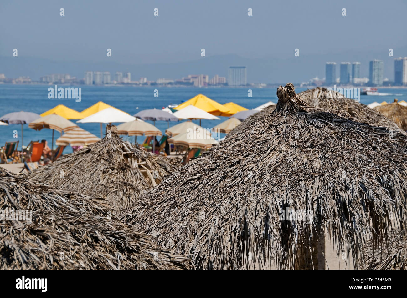 View of Puerto Vallarta and Banderas Bay over the tops of palapa and umbrella sources of shade on Los Muertos Beach. Stock Photo