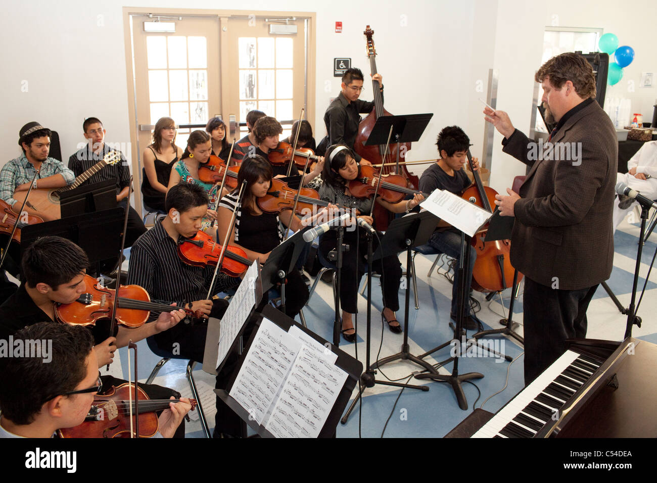 Students play stringed instruments, teacher conducts in orchestra class