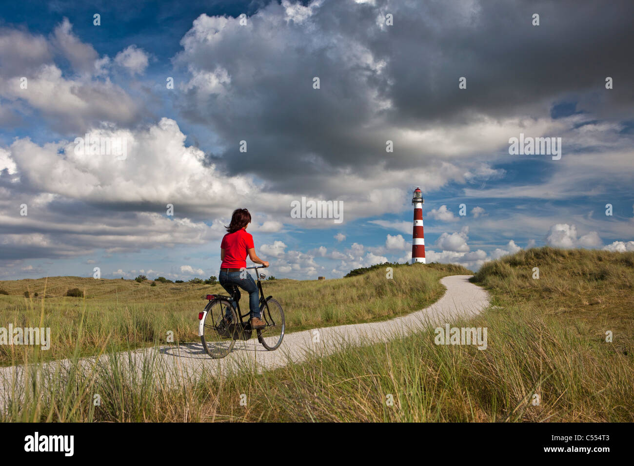 The Netherlands, Hollum, Ameland Island. Woman on bicycle on beach road. Lighthouse. Stock Photo