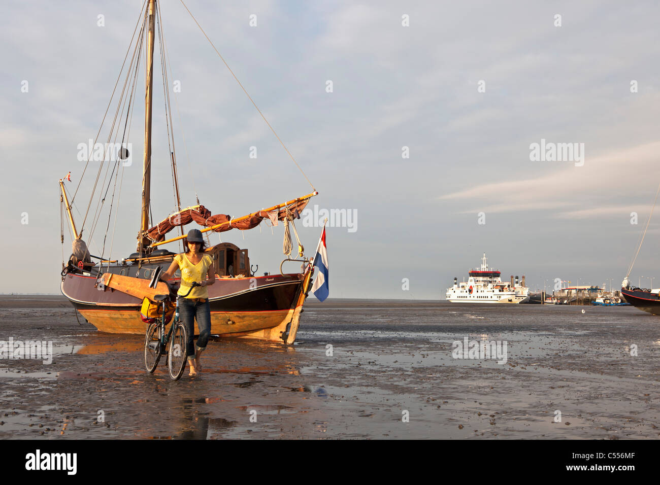 Nes, Ameland Island, Wadden Sea Islands. Sailing boat on mud flat in harbour. Woman and bicycle. Ferry boat to mainland. Stock Photo